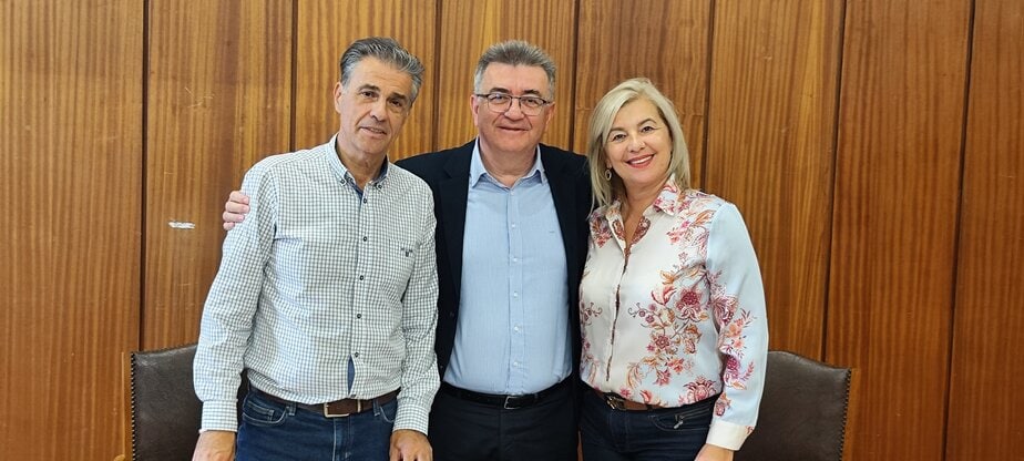 EKorinthos news team group photo with smiling professionals standing in front of wooden wall.
