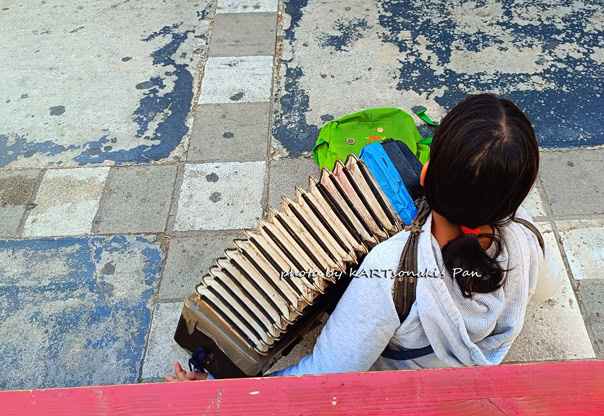 NET20241029_095537 Child sitting on the street with a musical instrument and backpack in Corinth, Greece.