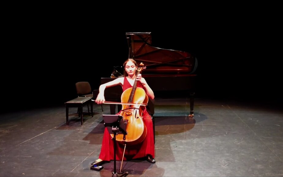 Elegant female cellist performing on stage with grand piano in the background.