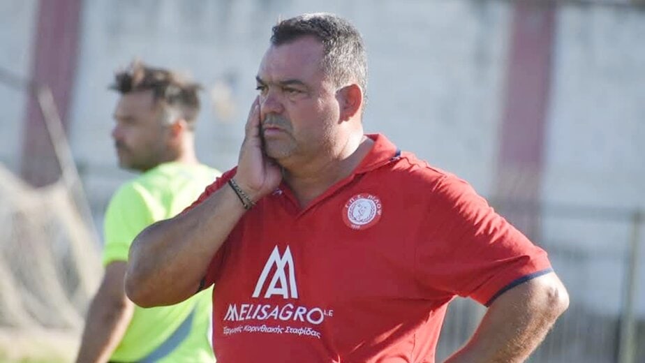 Frustrated football coach in red jersey during match, overlooking the field, with a concerned expression.